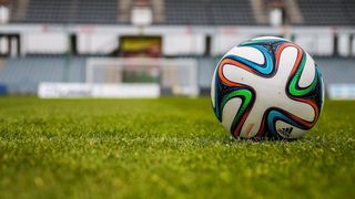 A soccer ball, adorned with colorful patterns, sits on a green field with an empty stadium in the background.