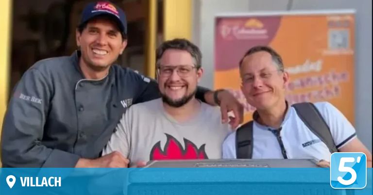 Three men smiling in front of a building. One has a chef's hat and the other two have glasses.