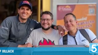 Three men smiling in front of a building. One has a chef's hat and the other two have glasses.