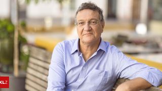 An older man wearing glasses and a light blue shirt sits on a wooden bench with a serious expression.