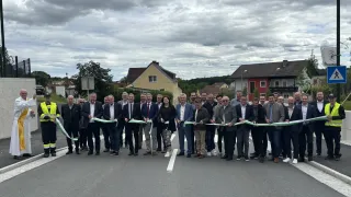 A group of people in suits and ties stand on the road, holding a ribbon for a ribbon-cutting ceremony.