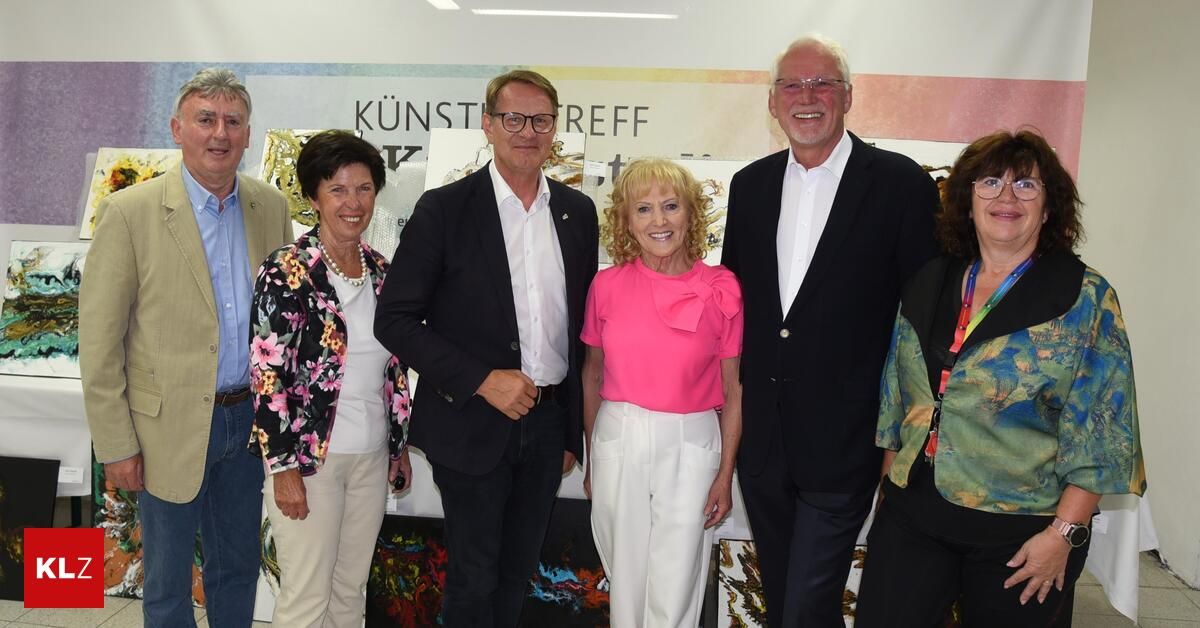A group of adults stands in front of a wall with artworks. The woman in the middle wears a pink blouse. The men wear dark suits. The woman on the left wears a floral jacket.
