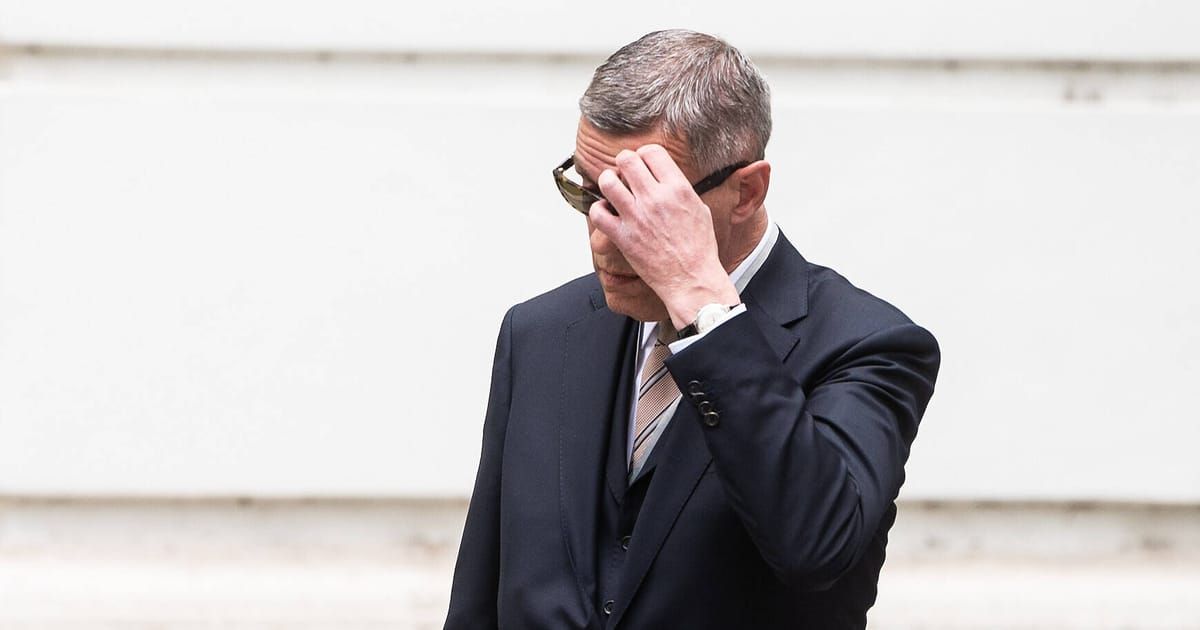 A man in formal attire, wearing a suit, tie, and glasses, is touching his face with his hand.