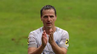 A man in a soccer uniform stands in the rain, clapping his hands. He wears a white shirt with yellow and black patches.