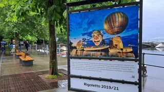 A billboard displays an image of a clown character from the opera Rigoletto, a hot air balloon, and a scenic view. The text describes Rigoletto as an oversized clown figure, emphasizing the emotional impact of the opera.