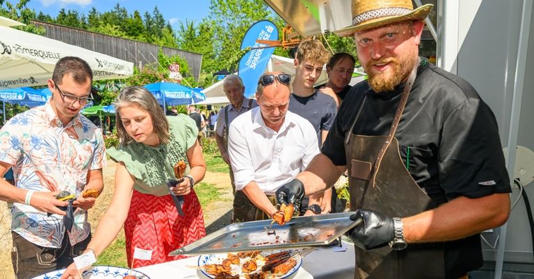 Ein Mann in Hut und Schürze kocht Essen auf einem Grill draußen. Eine Frau hält einen Spieß, und ein Mann schaut sie an. Andere Leute stehen um sie herum.
