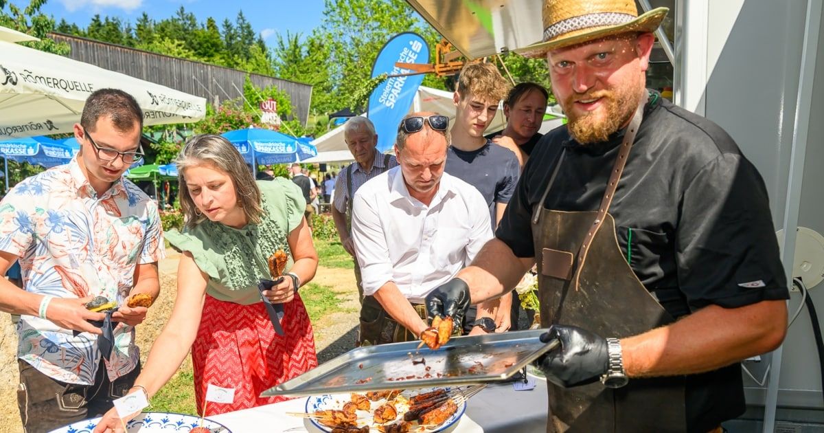 Ein Mann in Hut und Schürze kocht Essen auf einem Grill draußen. Eine Frau hält einen Spieß, und ein Mann schaut sie an. Andere Leute stehen um sie herum.