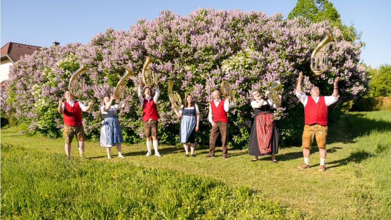 A group of people dressed in traditional Bavarian clothing stands in a field with flowering bushes behind them, holding brass instruments.