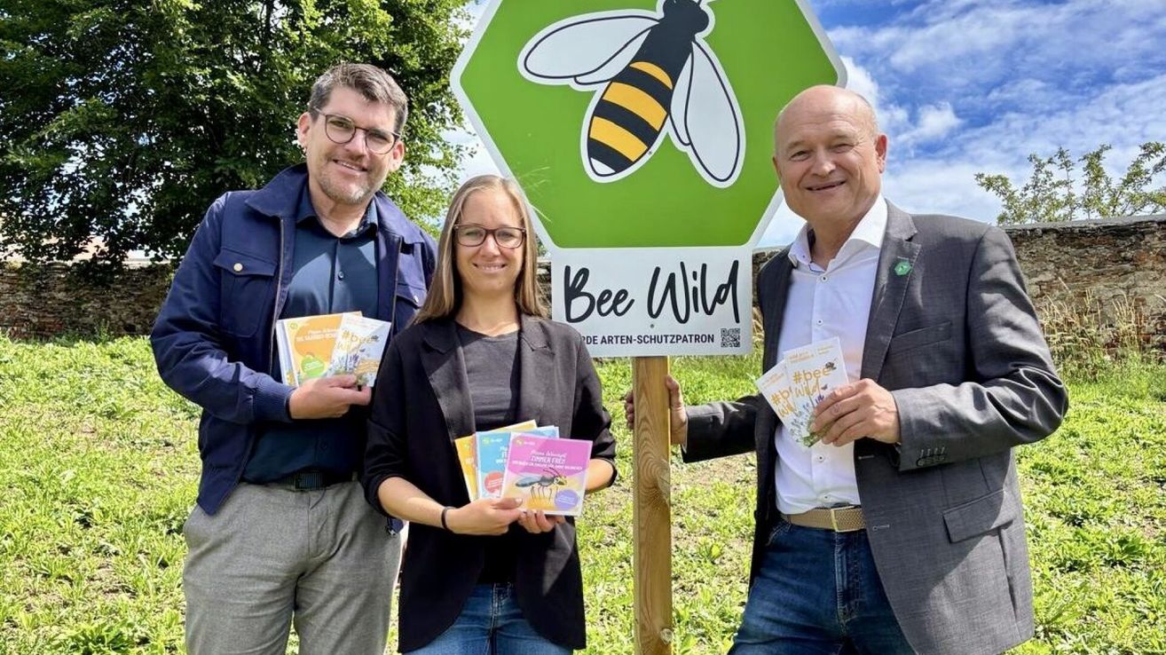 Drei Personen posieren mit Broschüren und lächeln in einem Park bei einem sechseckigen 'Bee Wild'-Schild mit einer Bienenillustration.