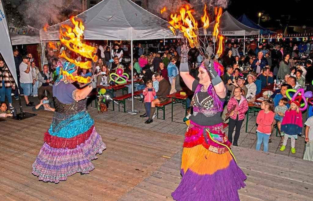 Two performers juggle flaming batons in front of an audience. Both wear colorful, elaborate costumes. Tables and tents are set up in the background.