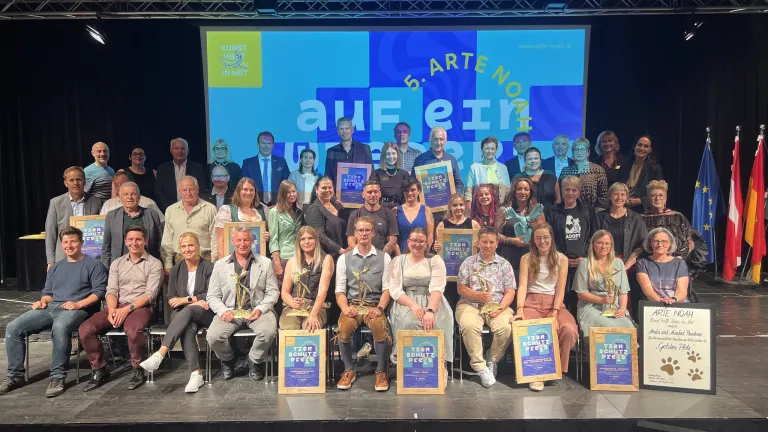 Eine Gruppe von Menschen mit Pokalen posiert auf einer Bühne, wobei ein großer Bildschirm im Hintergrund 'Kunst Hilft in Not' und '5. Arte Nothilfe' anzeigt.