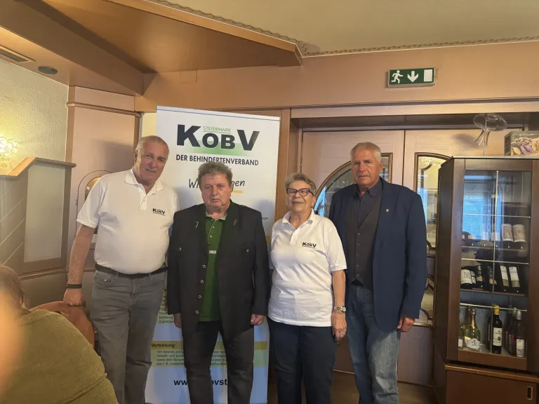 Vier Personen stehen vor einem Banner mit den Buchstaben KOBV, möglicherweise bei einer Veranstaltung. Ein Mann links trägt ein weißes Poloshirt, während ein anderer Mann in der Mitte einen schwarzen Anzug trägt. Die Frau rechts trägt ein weißes Shirt. Ein Mann rechts trägt einen Anzug. Dahinter ist eine Tür mit einem Ausgangsschild zu sehen.