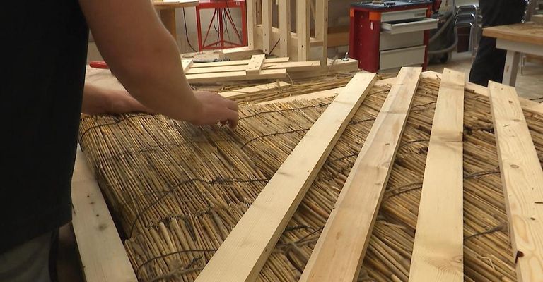 Eine Person arbeitet an einem Strohdach mit Holzbrettern und Bambus. Ein roter Werkzeugschrank und eine Holzstruktur sind im Hintergrund.