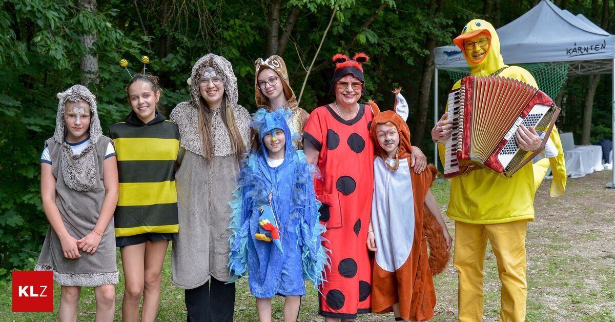 A group of people dressed in various costumes are posing for a picture. The costumes include a ladybug, a fox, a bird, a bee, an owl, and a yellow duck. The background features trees and a canopy.