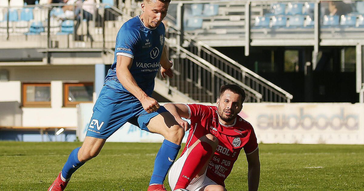 Two soccer players are on the field, one in blue and the other in red. The player in blue is attempting to steal the ball while the player in red is sitting on the ground.