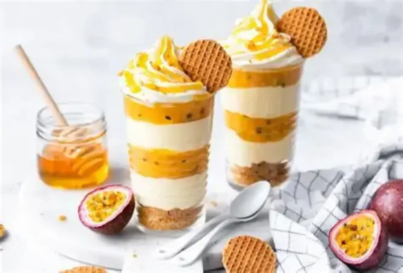Zwei Dessertbecher mit Schlagsahne und Waffeln, geschichtet mit einer Mischung aus Orange und Passionsfrucht, begleitet von einem Glas Honig, einem Löffel und einer Waffel auf einer weißen Oberfläche.
