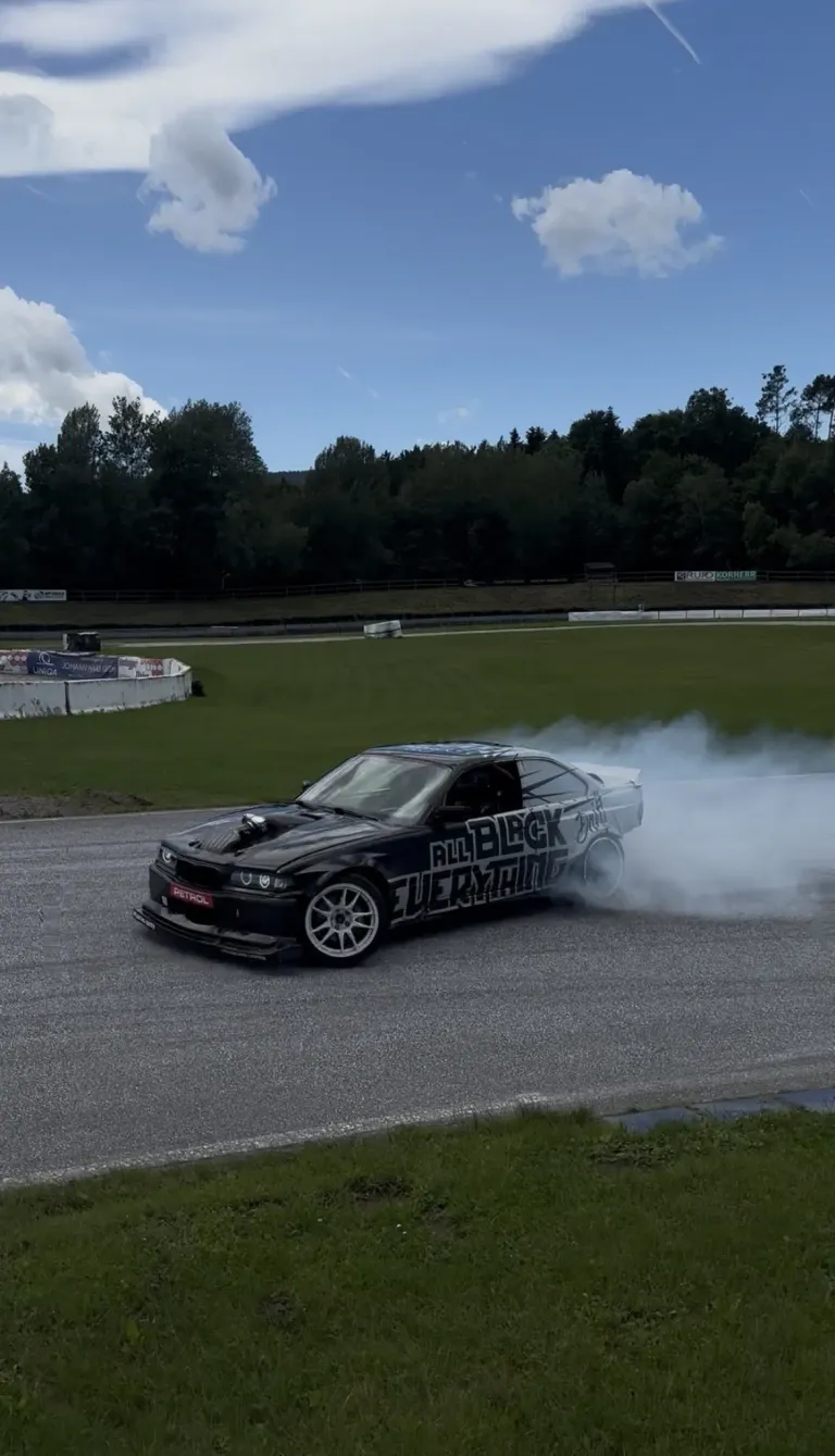Ein schwarzer Wagen mit der Aufschrift 'All Black Everything' fährt auf einer Rennstrecke mit Rauch aus den Reifen. Im Hintergrund befindet sich eine Wiese mit Bäumen.