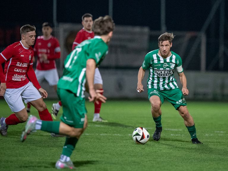 Fußballspieler in grünen und roten Trikots auf einem Feld. Der Spieler in Grün verfolgt den Ball, während andere zusehen.