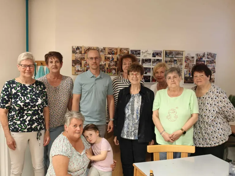 Eine Gruppe älterer Frauen und Männer steht lächelnd für ein Foto in einem Raum zusammen. Ein kleines Mädchen sitzt auf dem Schoß einer Frau. Dahinter befinden sich mehrere gerahmte Bilder an einer weißen Wand.
