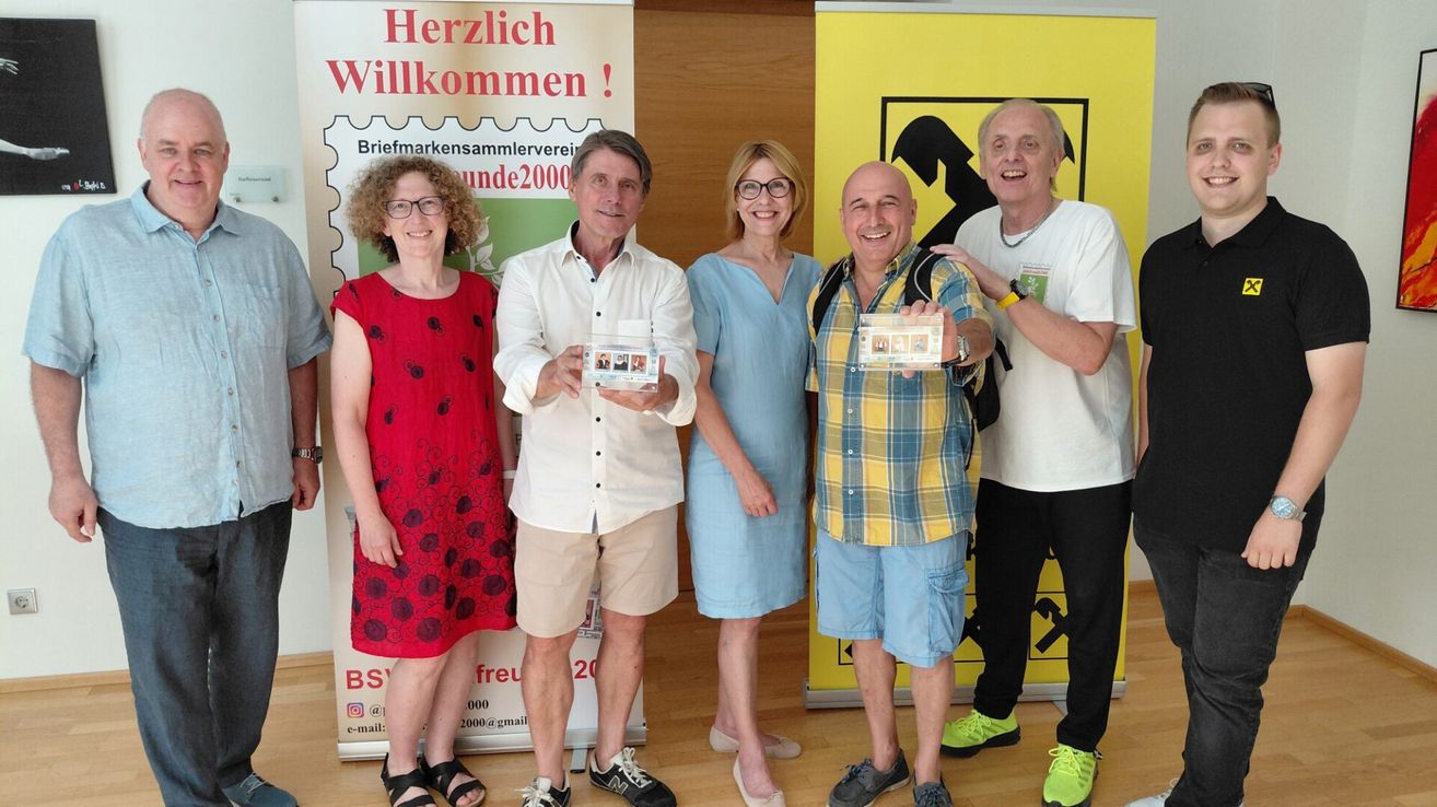 Fünf Menschen stehen vor einem Schild mit der Aufschrift Herzlich Willkommen. Zwei Männer und drei Frauen halten Karten mit Bildern von Personen in die Luft.