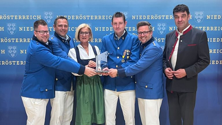 Fünf Personen in Uniform stehen zusammen und halten eine Trophäe, mit einem blauen Hintergrund, der Niederosterreich steht.