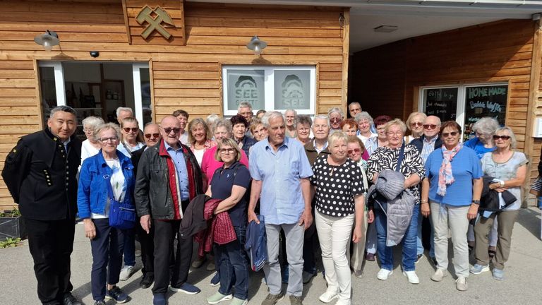 Eine Gruppe älterer Personen versammelt sich vor einem hölzernen Gebäude mit dem Wort SEE. Sie lächeln und posieren für ein Foto.