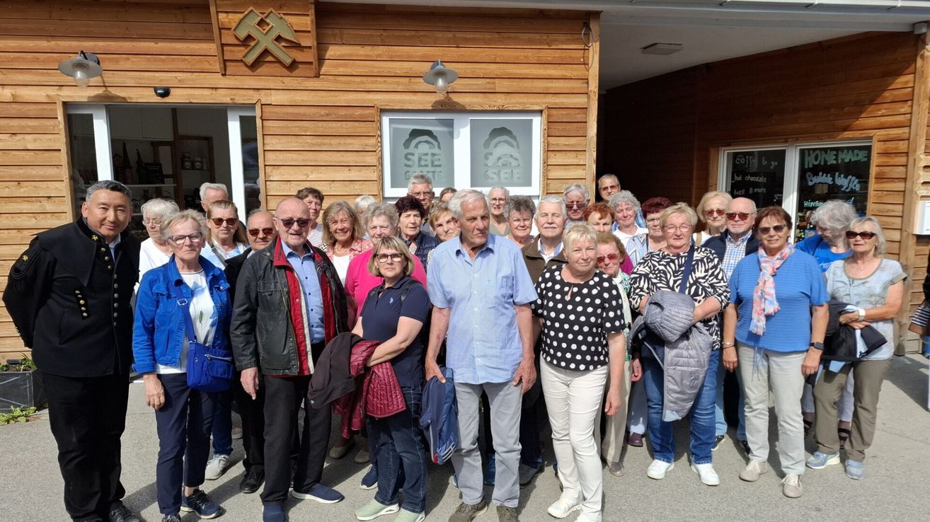 Eine Gruppe älterer Personen versammelt sich vor einem hölzernen Gebäude mit dem Wort SEE. Sie lächeln und posieren für ein Foto.