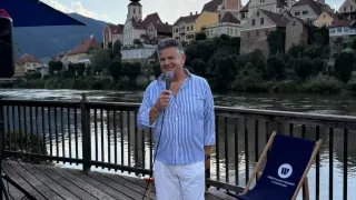Ein Mann in einem gestreiften Hemd und weißen Shorts steht auf einem Deck, hält ein Mikrofon und hat einen Fluss und eine Stadt hinter sich.