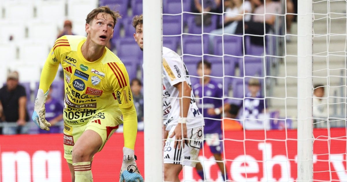 Ein Fußballspieler in einem gelben Trikot steht in der Nähe eines Tores, mit einem anderen Spieler in einem weißen Trikot dahinter. Zuschauer sitzen auf den Tribünen.