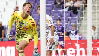 Ein Fußballspieler in einem gelben Trikot steht in der Nähe eines Tores, mit einem anderen Spieler in einem weißen Trikot dahinter. Zuschauer sitzen auf den Tribünen.