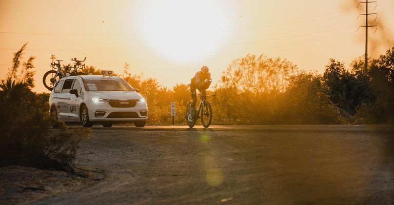 Ein Radfahrer fährt auf einer Straße, während ein weißer Wagen mit Radar und Lichtern auf dem Dach dahinterher fährt. Der Sonnenuntergang ist im Hintergrund zu sehen.