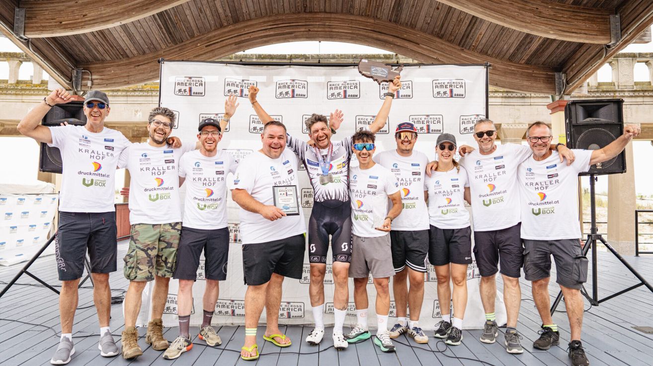 Eine Gruppe von Radfahrern posiert für ein Foto auf der Bühne, alle tragen passende weiße Shirts mit Logos und Sonnenbrillen. Ein Mann hält eine Trophäe. Dahinter steht ein Banner mit der Aufschrift 'Race Across America.'