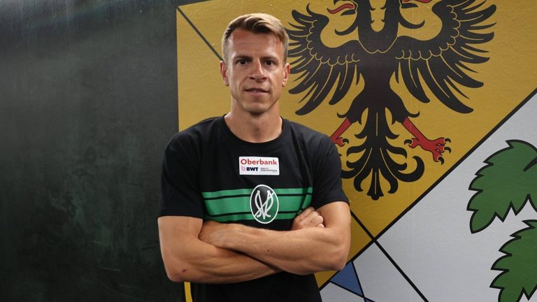 Ein Mann steht mit verschränkten Armen vor einer gelben Wand mit einem Adler und einem Logo. Er trägt ein schwarzes Oberbank-Shirt.
