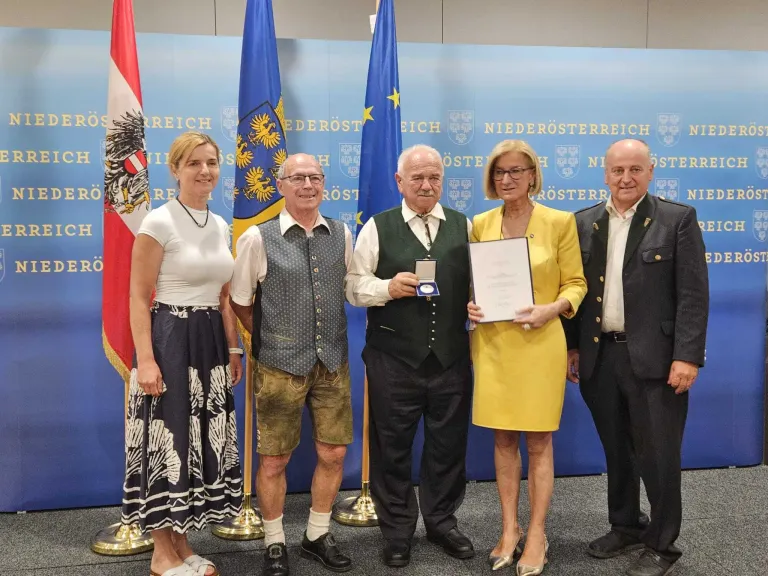 Fünf Personen stehen vor einem Hintergrund mit Fahnen. Eine Frau hält ein Zertifikat und eine Medaille. Ein anderer Mann hält eine Medaille. Sie scheinen einen formellen Anlass zu feiern.