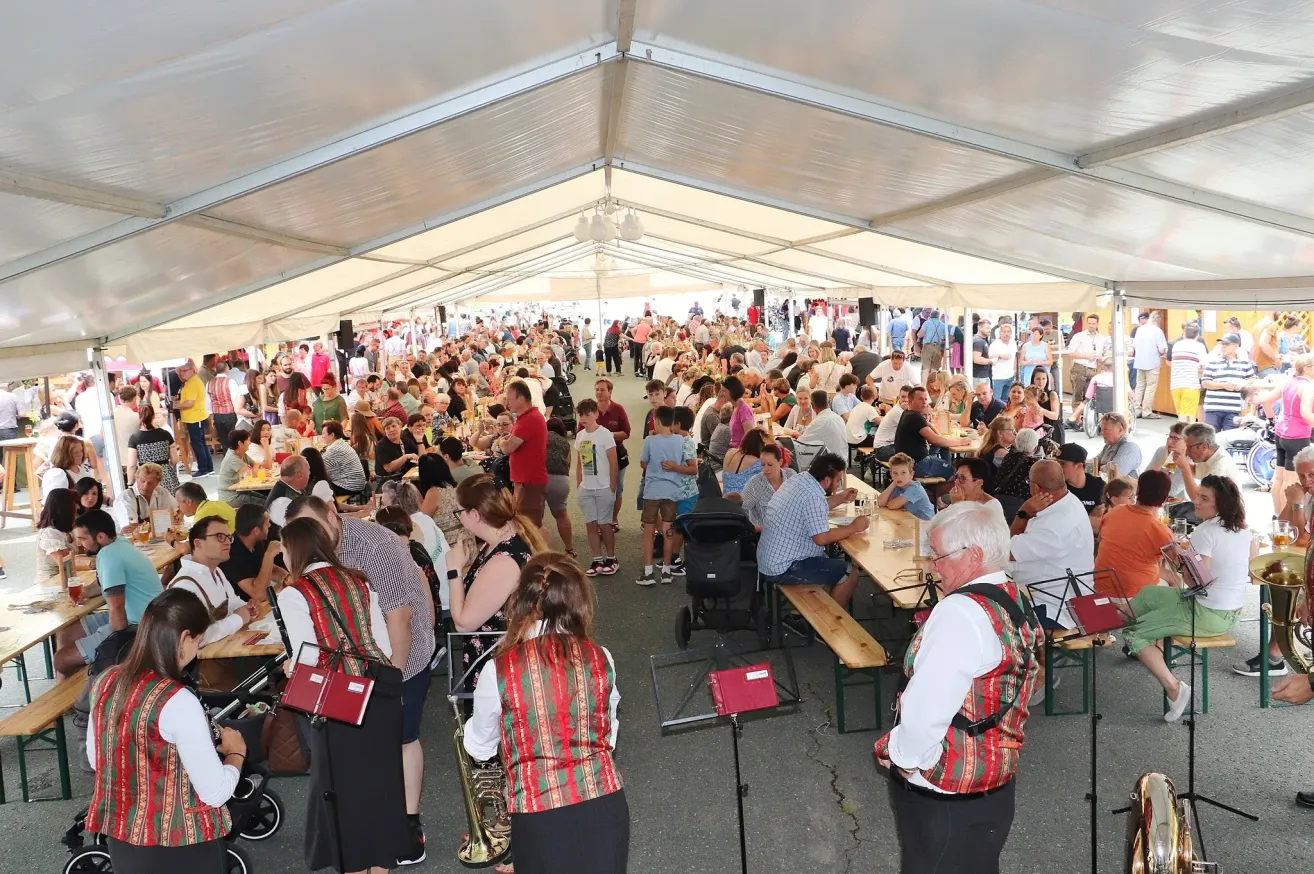 Eine große Menschenmenge speist unter einem Zelt auf einem Festival. Viele tragen traditionelle Kleidung. Ein Musiker steht mit einem Blechinstrument, und die Tische sind mit Essen gefüllt.