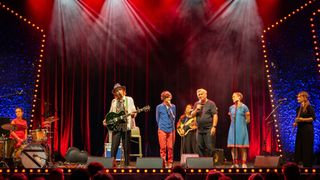 Eine Band tritt auf der Bühne unter roter Beleuchtung auf. Eine Person spielt Gitarre, während die anderen Mikrofone halten. Ein Publikum beobachtet von vorne.