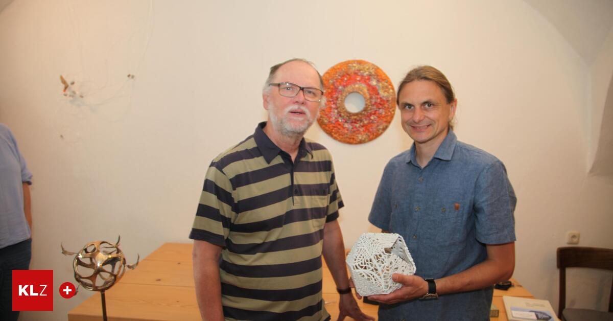 Zwei Männer stehen vor einer Wand mit bunter Kunst. Einer hält eine weiße geometrische Skulptur. Sie lächeln beide.