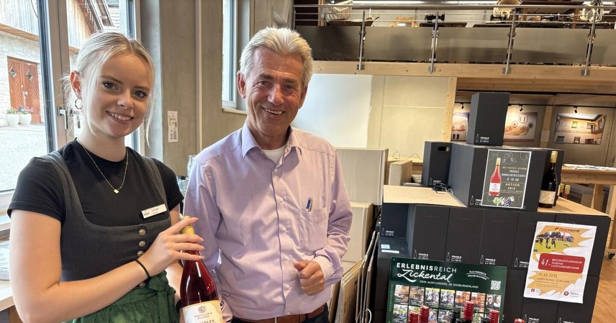Ein Mann und eine Frau stehen zusammen in einem Raum mit Regalen voller Weinflaschen. Die Frau hält eine Flasche Wein, und der Mann lächelt. Hinter ihnen befinden sich mehrere Kisten mit Wein.
