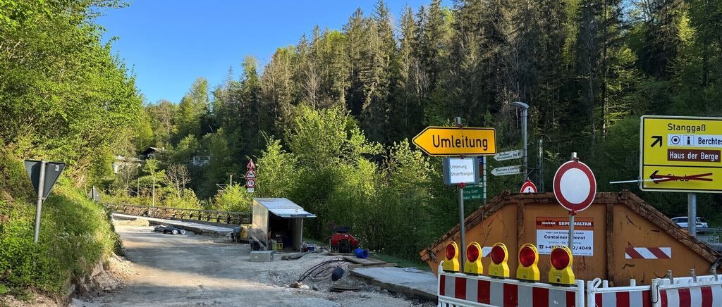 Baustelle mit einem gelben Schild, das eine Umleitung anzeigt. Es gibt Verkehrskegel und ein rotes und weißes Schild. Das Gebiet ist von Bäumen und Büschen umgeben.