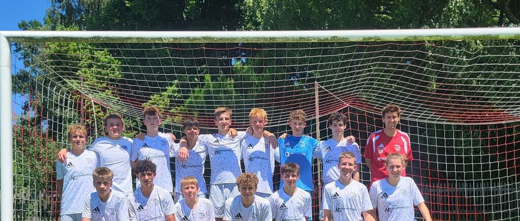 Eine Gruppe junger männlicher Fußballspieler in weißen Trikots posiert für ein Foto vor einem Torpfosten mit Bäumen im Hintergrund.