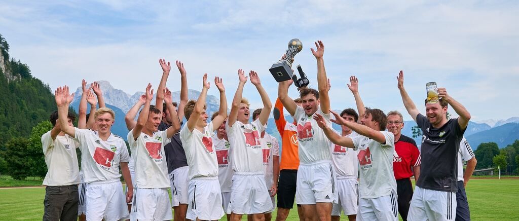 Ein Fußballteam feiert ihren Sieg auf einem Feld mit Bergen im Hintergrund. Sie halten einen Pokal hoch, und alle lächeln.