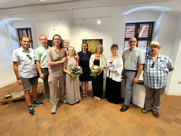 Eine Gruppe von Menschen steht in einem Raum mit einer Frau in der Mitte, die einen Blumenstrauß hält. Einige tragen eine Brille und Turnschuhe. Eine Frau hält ein Papier, und ein Mann trägt einen Hut. Der Raum hat weiße Wände, eine weiße Decke und geflieste Böden.