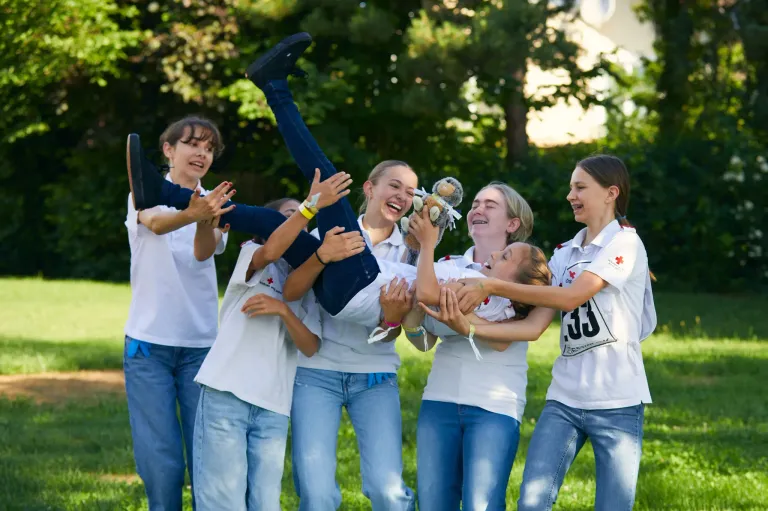 Eine Gruppe junger Mädchen in weißen Hemden und Jeans trägt ein Kind auf einem Rasenfeld. Sie alle lächeln und scheinen Spaß zu haben.