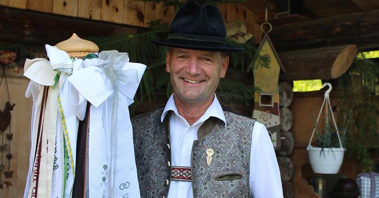 Ein lächelnder Mann in traditioneller Kleidung steht neben einer dekorativen Schleife und einem Vogelhaus.