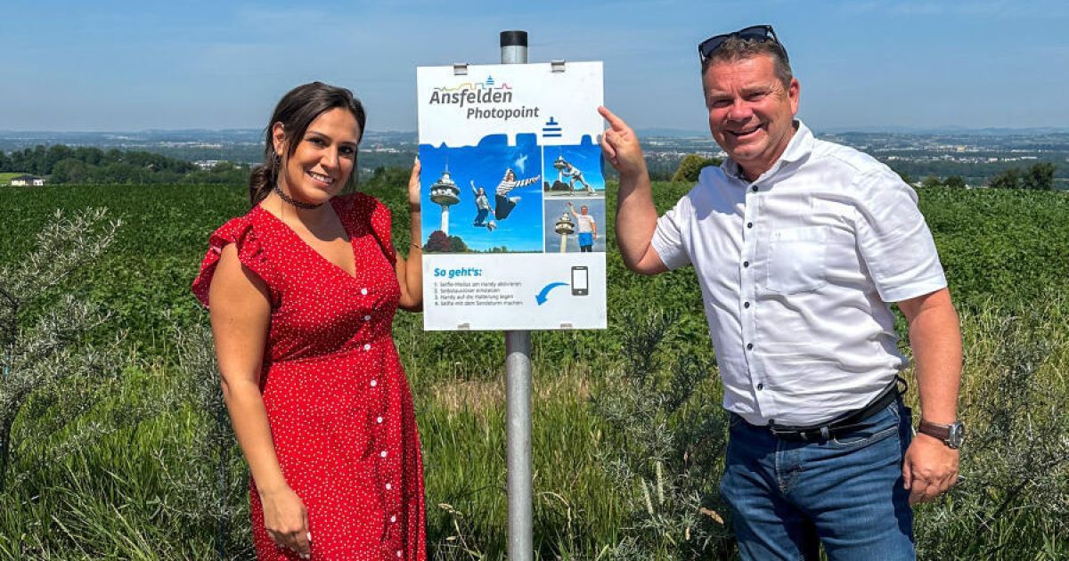 Ein Paar steht neben einem Fotopunkt-Schild in einem Grasfeld, der Mann zeigt lächelnd auf das Schild.