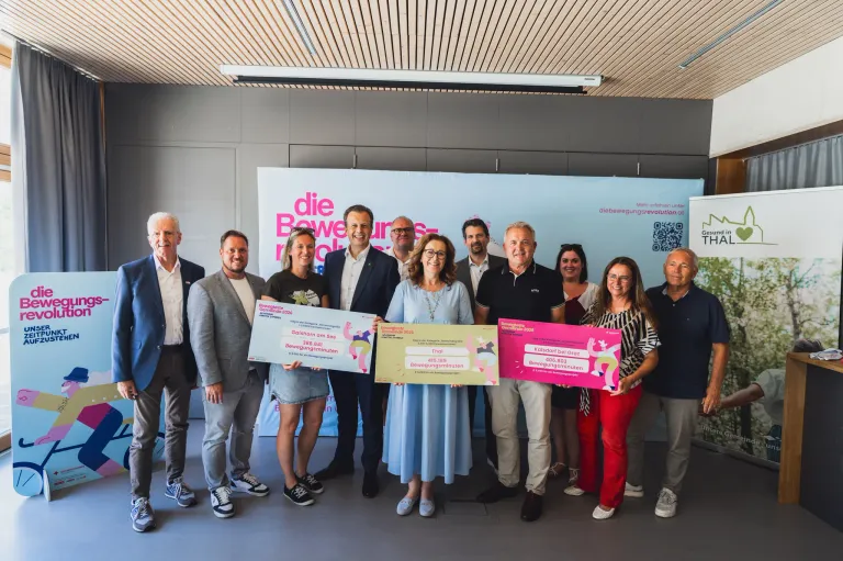 A group of individuals stands together, holding large checks, in front of a banner with pink text and a QR code. They appear to be celebrating a significant achievement.