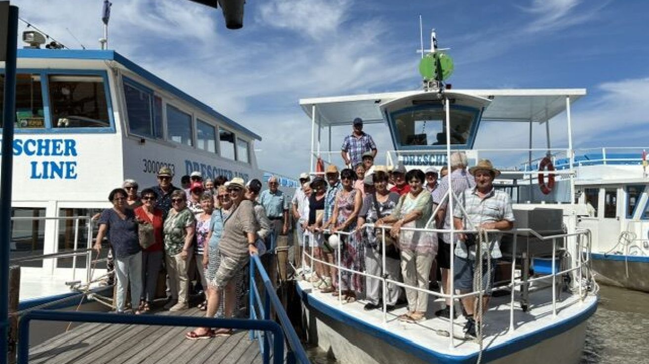 Eine Gruppe von Menschen steht auf einem Boot, einige tragen Hüte und Brillen. Eine Person hält eine Reling. Das Boot heißt DRESSCHER und liegt an einem Pier.