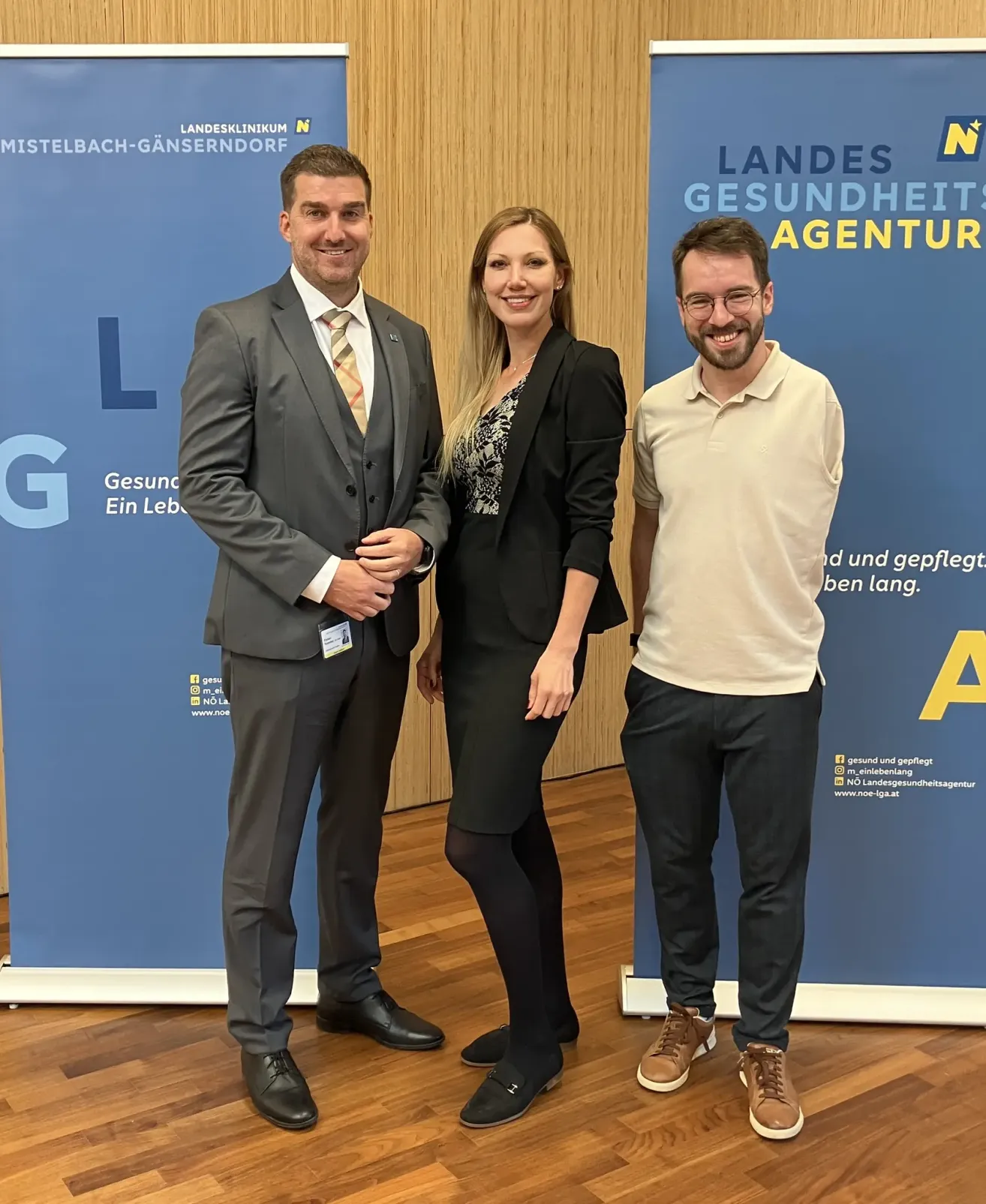 Drei Personen, zwei Männer und eine Frau, stehen nebeneinander vor einem blauen Hintergrund. Sie lächeln und scheinen für ein Foto zu posieren.