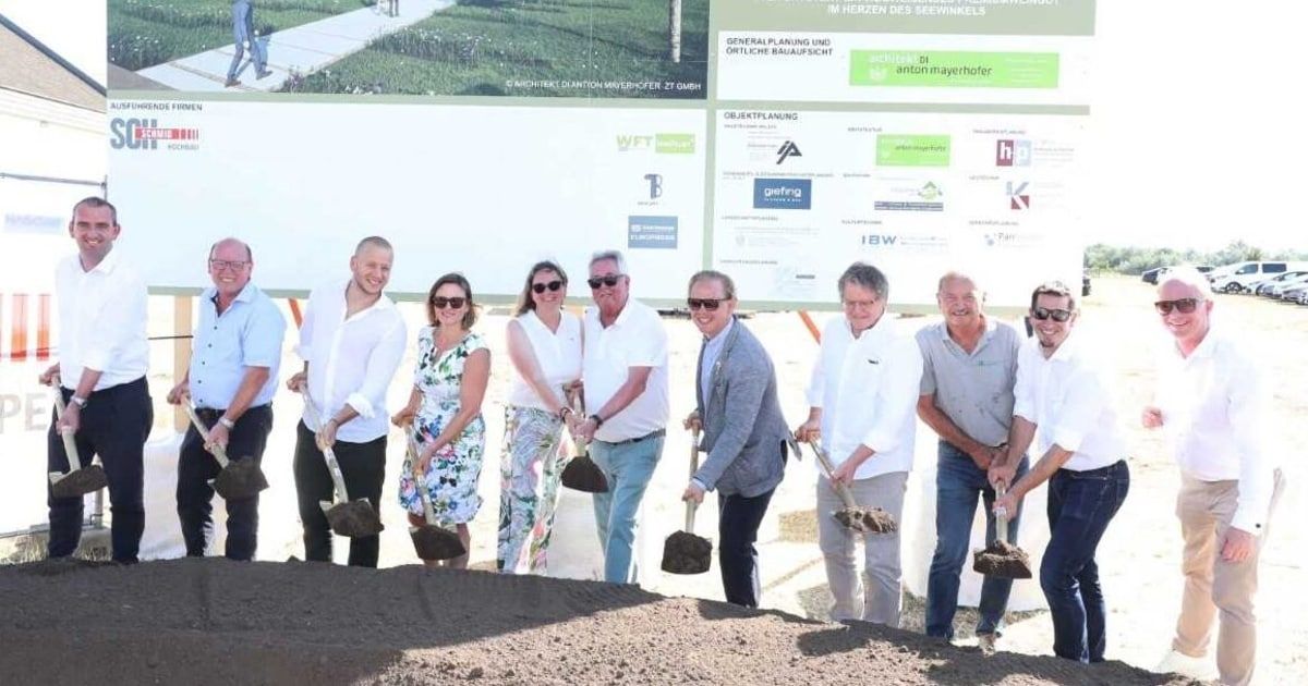 Eine Gruppe von Menschen mit Schaufeln, Sonnenbrillen und Turnschuhen steht auf einem Haufen Erde vor einem großen Schild.