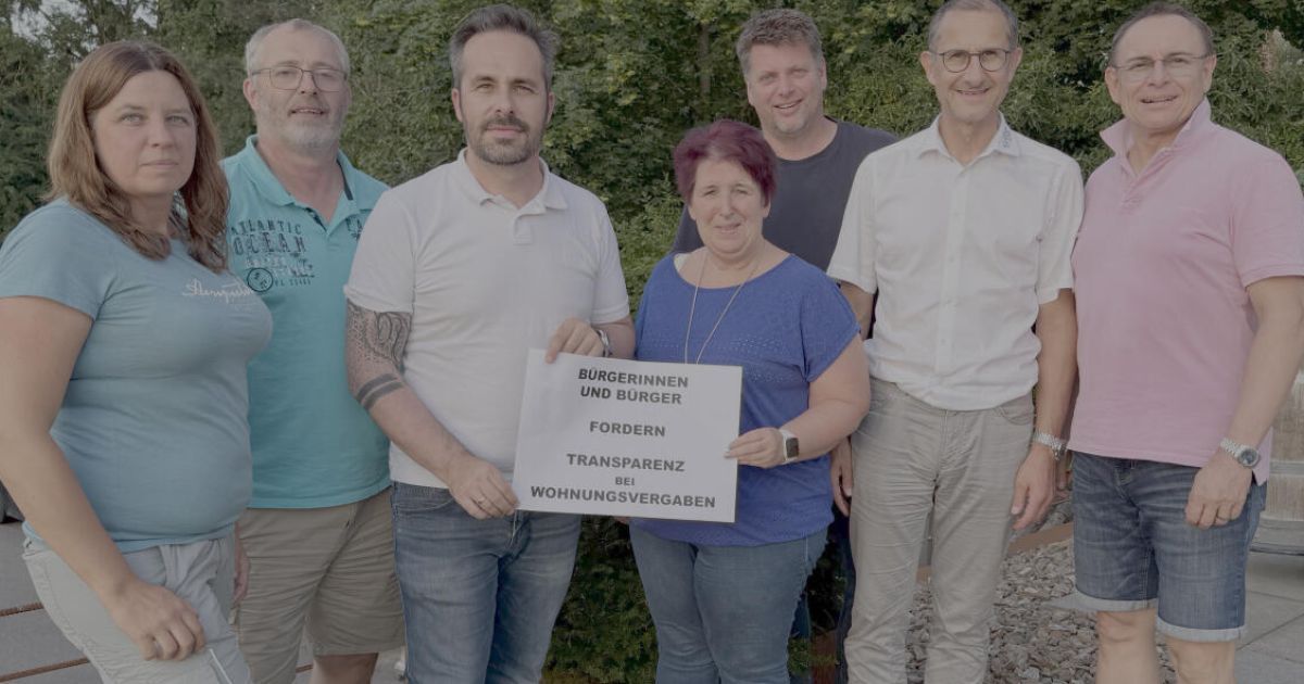Fünf Erwachsene stehen lächelnd für ein Foto draußen. Der Mann links hat ein Tattoo. Der Mann in der Mitte hält ein Schild mit der Aufschrift 'BURGERN UND BURGER FORDERN TRANSPARENZ BEI WOHNUNGSVERGABEN.' Die Frau in der Mitte trägt eine Halskette.